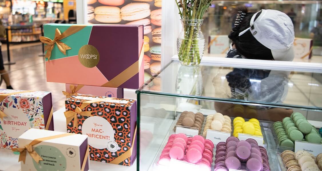 A Woops! kiosk showing macaron boxes and a counter full of French macarons. 