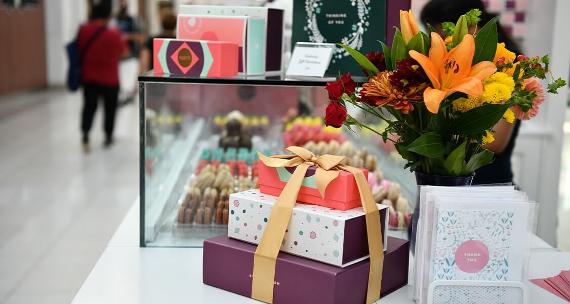 A Woops! kiosk counter with French macaron boxes, a macaron display, and a vase with flowers. 