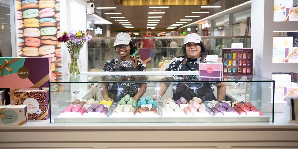 Two smiling women are behind a Woops! kiosk counter that’s full of French macarons and macaron boxes.