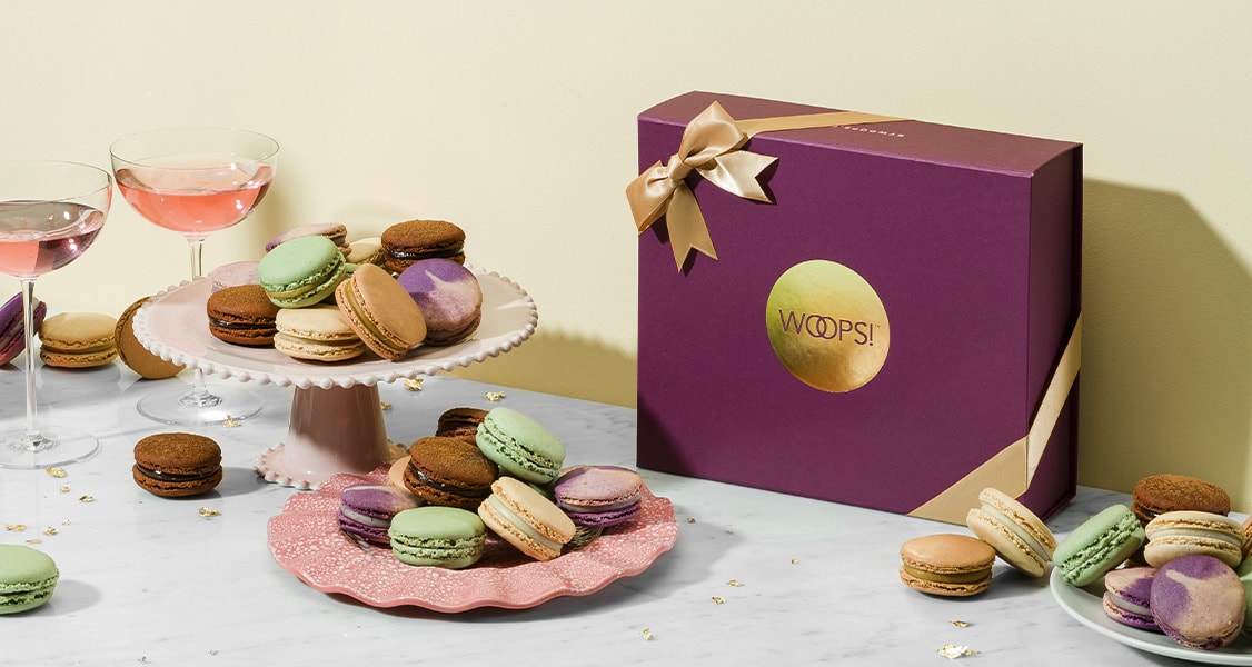 A Woops! box of French macarons is surrounded by plates full of French macarons and cups of champagne.