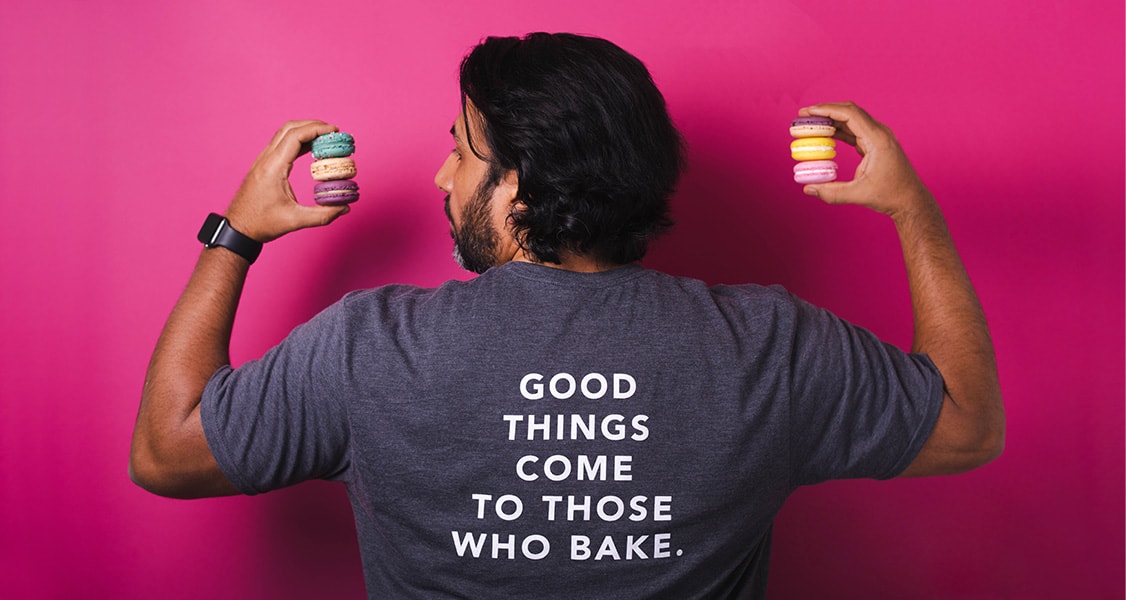 A black-haired man is holding assorted French macarons in his hands. He has a T-shirt that says “good things come to those who bake” on his back.