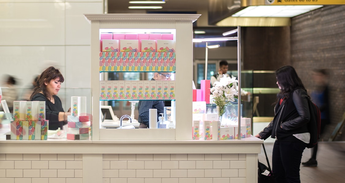 A white Woops! Macarons & Gifting kiosk is full of macaron boxes and macaron pyramids.