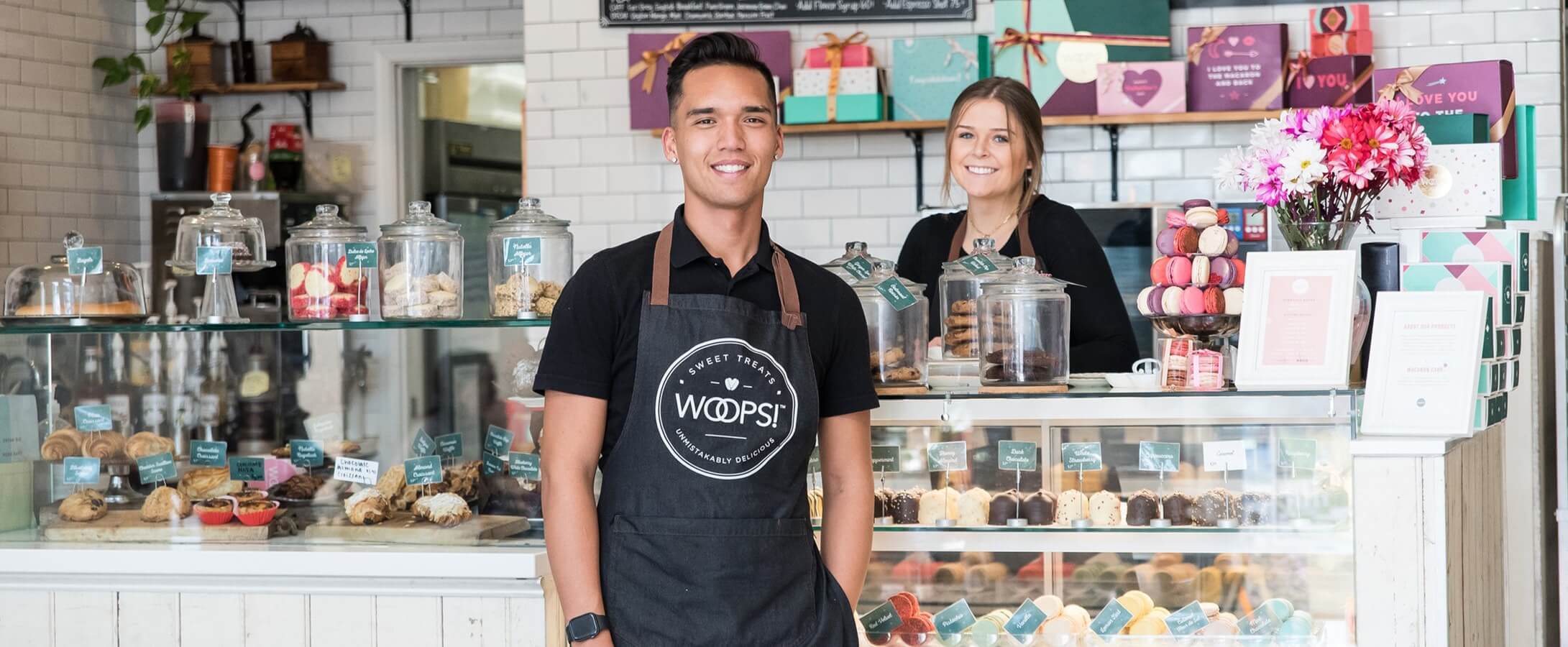 A smiling man and a woman wearing Woops! aprons are surrounded by macarons, macaron boxes, a display case with macarons, and cookies.