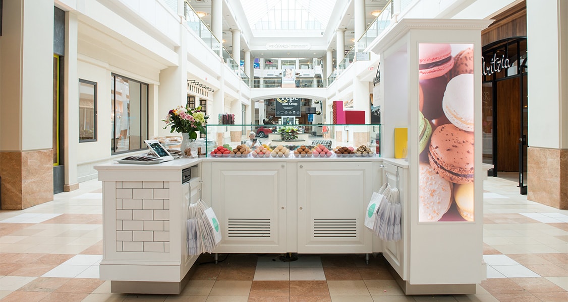 A white Woops! mall kiosk has a big banner displaying macarons and a counter full of assorted French macarons