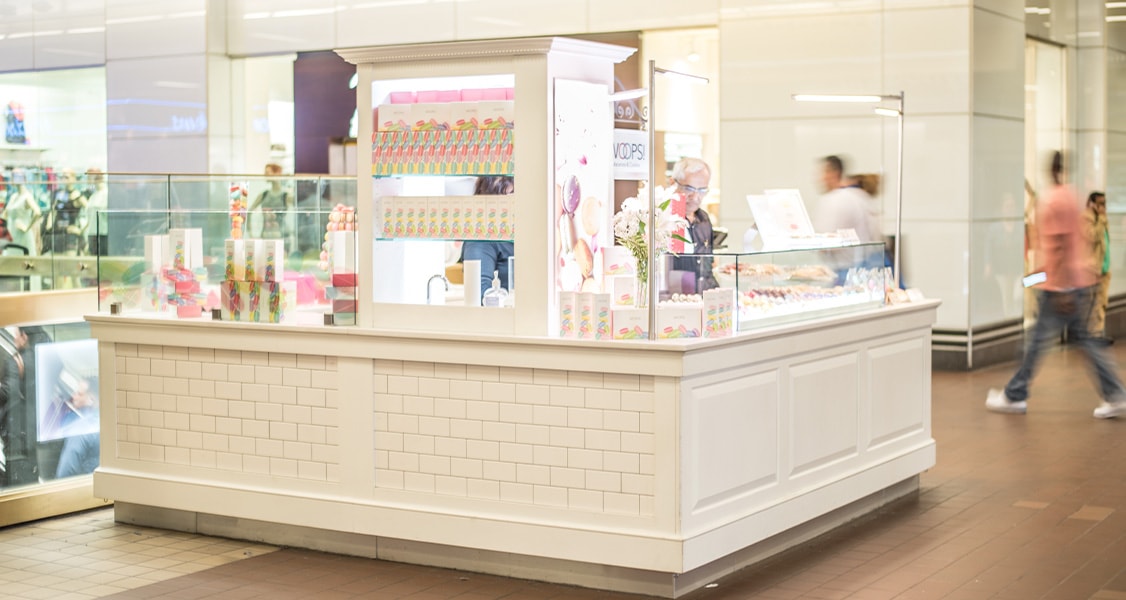 A WOOPS! Macarons & Gifts white kiosk contains assorted French macarons and gift boxes.