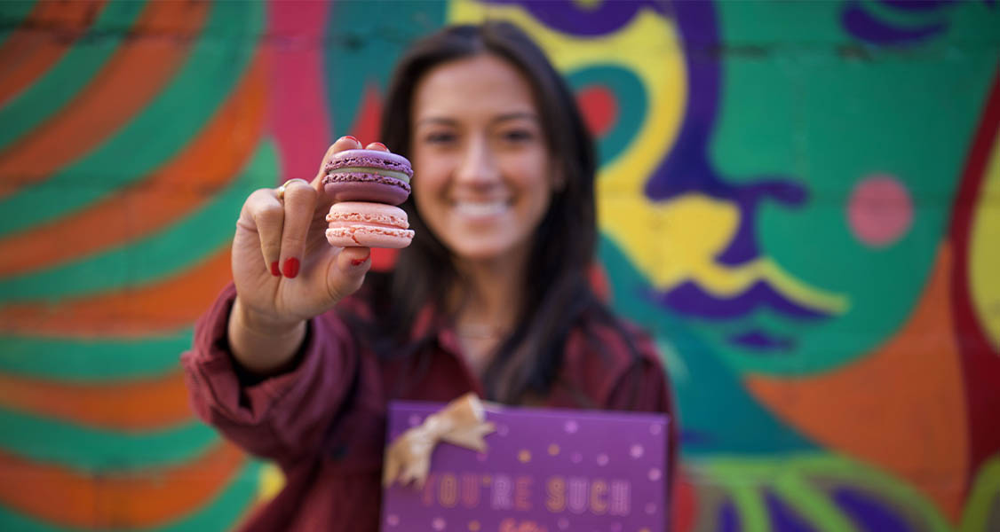 A smiling woman is holding two French macarons in one hand and a WOOPS! Macarons & Gifts purple macaron box in the other. 
