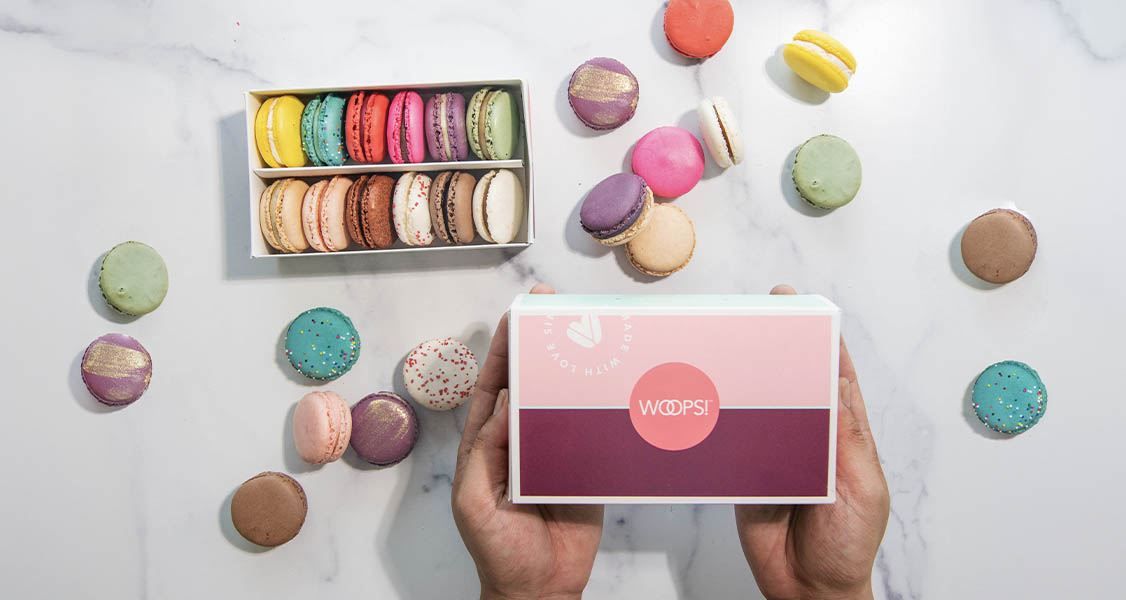 A pair of hands are holding a pink box of Woops! French macarons. A box full of assorted macarons serves as background.