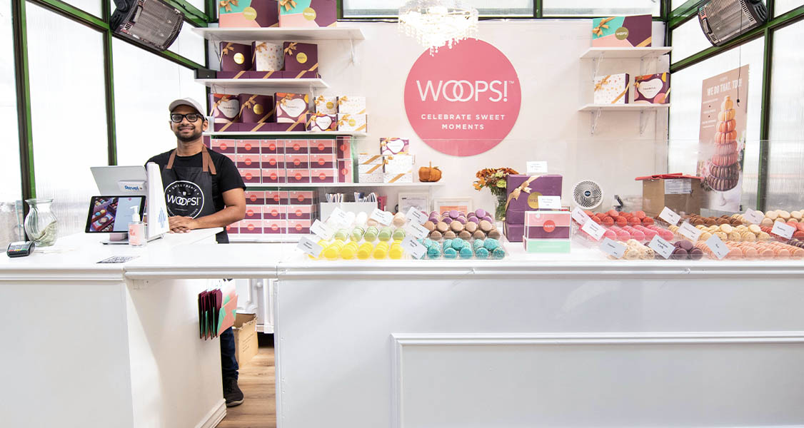 A smiling man is standing behind a Woops! counter that’s full of assorted French macarons and French macaron boxes.