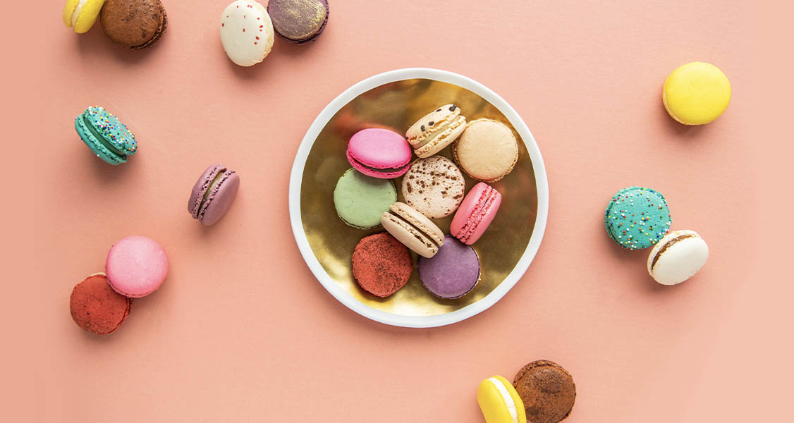 A plate full of assorted macarons is surrounded by more macarons.