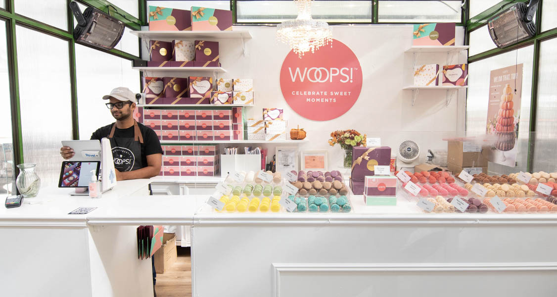 A smiling man is standing behind a WOOPS! Macarons & Gifts counter that’s full of assorted macarons and macaron boxes.