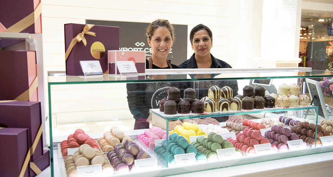 Two smiling women are standing behind a WOOPS! Counter that’s full of assorted macarons and cremebellos. 
