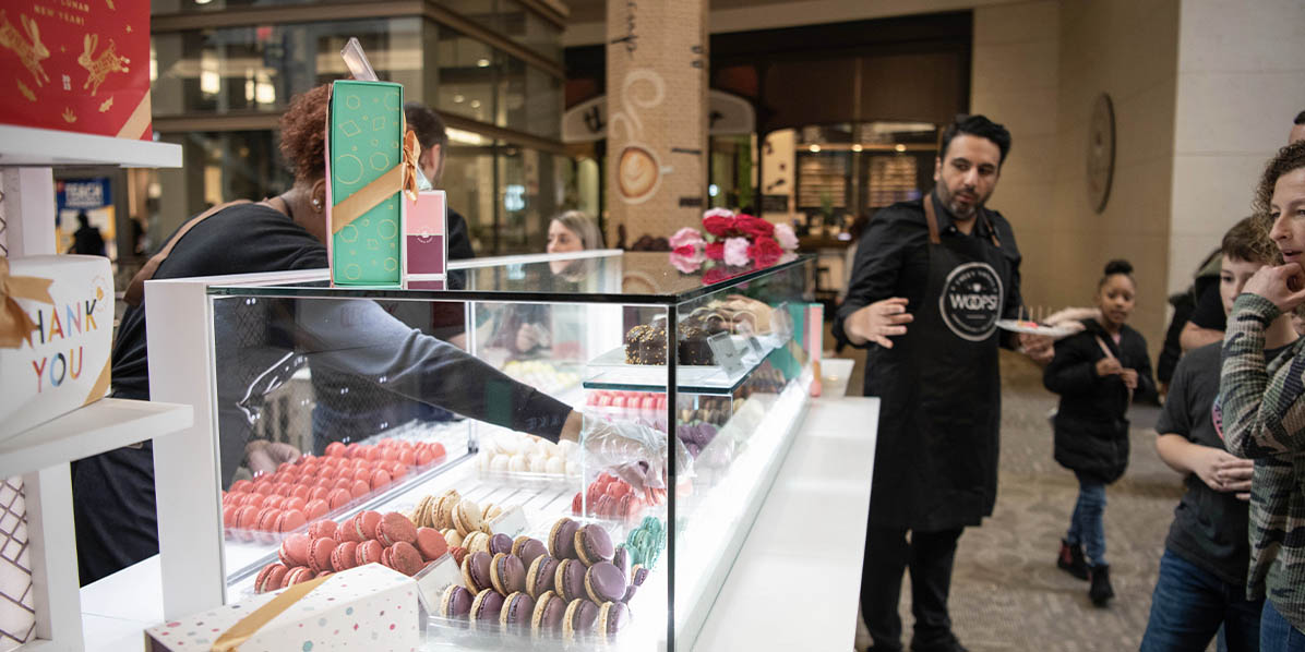 A man is standing in front of a WOOPS! Macarons & Gifts kiosk that’s full of assorted macarons and macaron boxes.