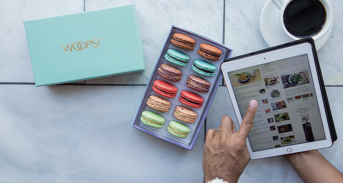 an open box of macarons, a cup of coffee, and someone using a finger to scroll on an iPad