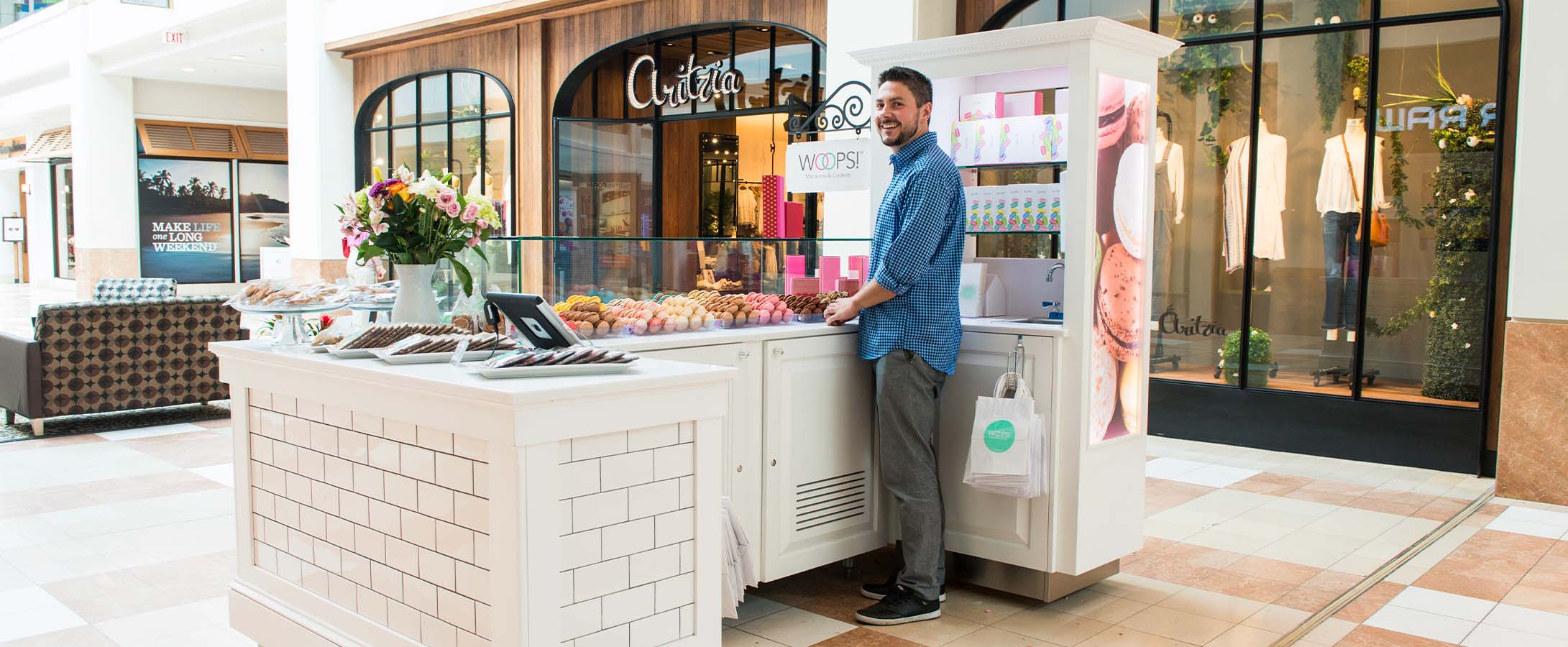 A smiling man is standing behind a Woops! kiosk counter that’s full of assorted French macarons and colorful macaron boxes.