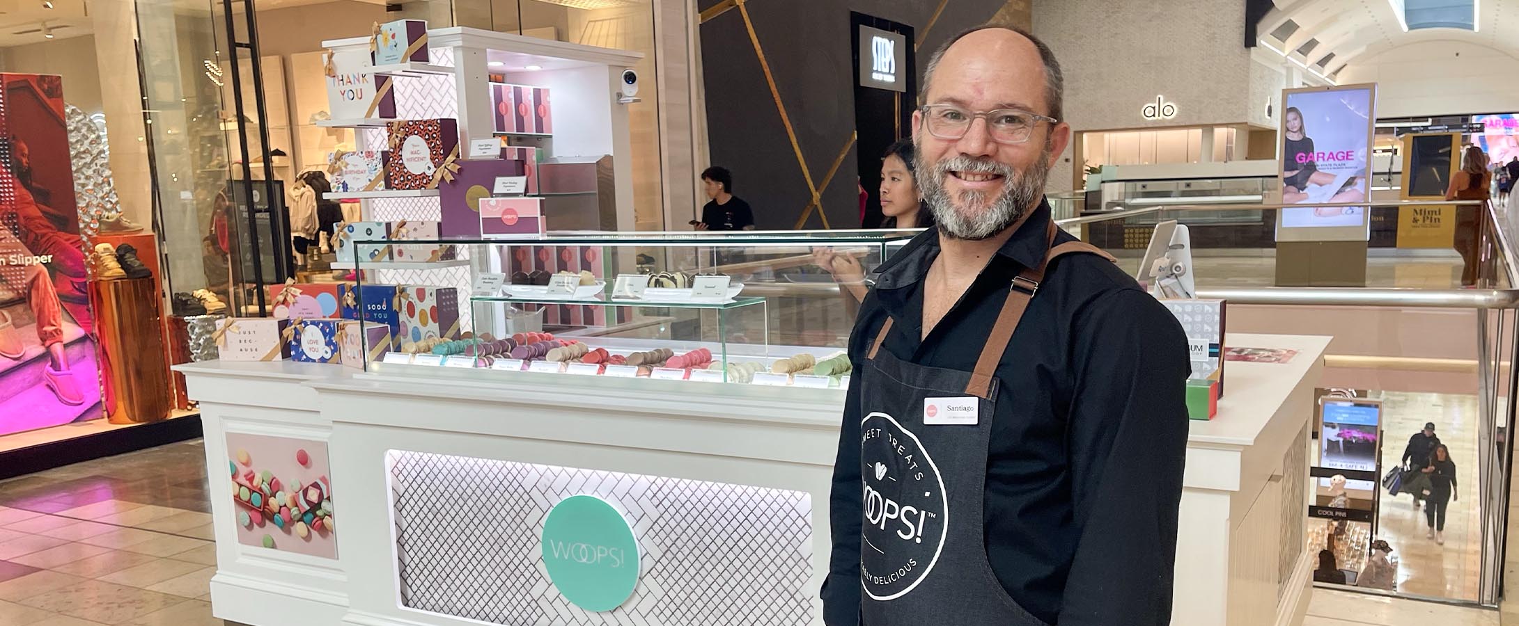 A smiling man with a Woops! Apron stands in front of Woops! Macarons & Gifts macaron kiosk.