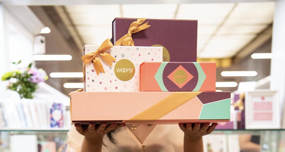 A woman is carrying multiple Woops! Macarons & Gifts gift bags and gift boxes.