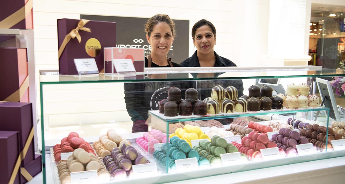 Two smiling women are standing behind a Woops! kiosk counter full of assorted macarons and macaron boxes. 