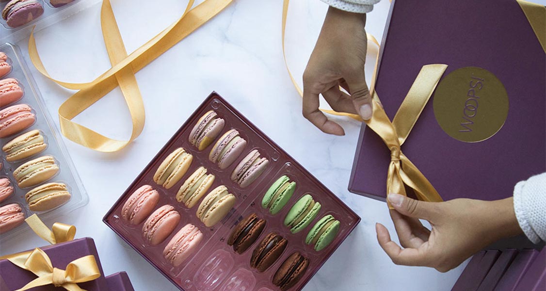 Two female hands are tying a golden-colored ribbon on a purple macaron box. All around are French macaron boxes and trays full of macarons.