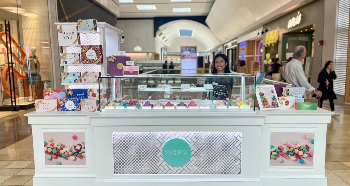 A smiling woman is standing behind a Woops! Macaron kiosk full of assorted macarons and macaron boxes. 
