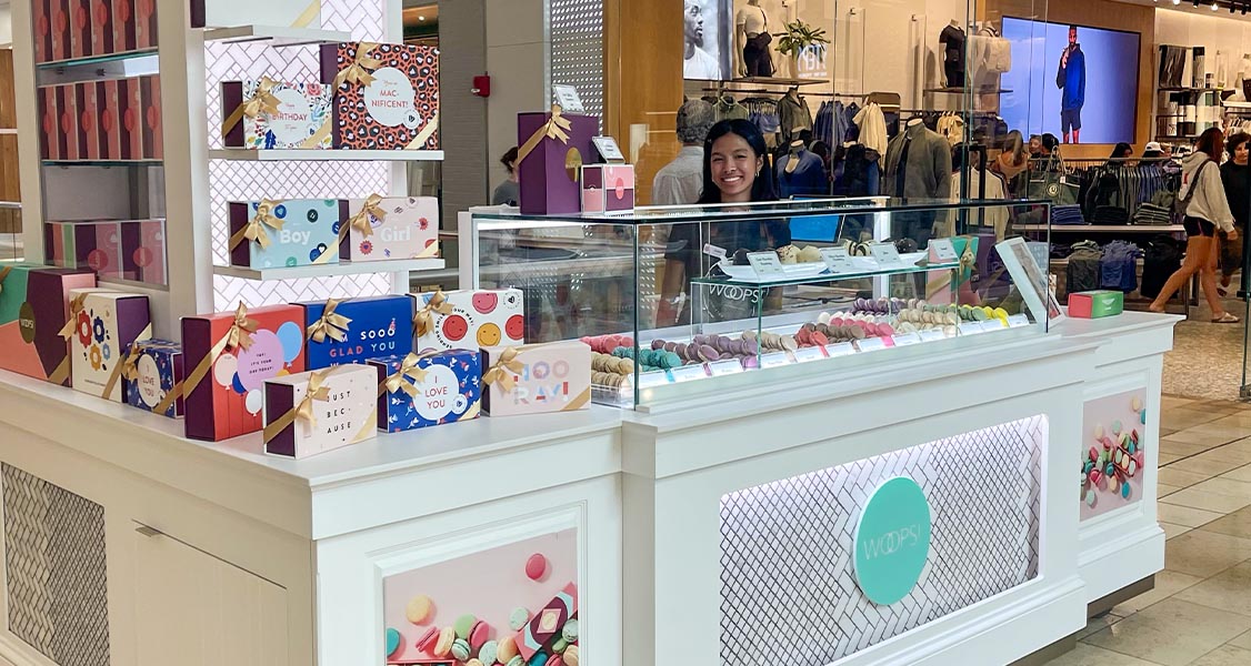 A smiling woman is standing behind a Woops! Macaron kiosk. 