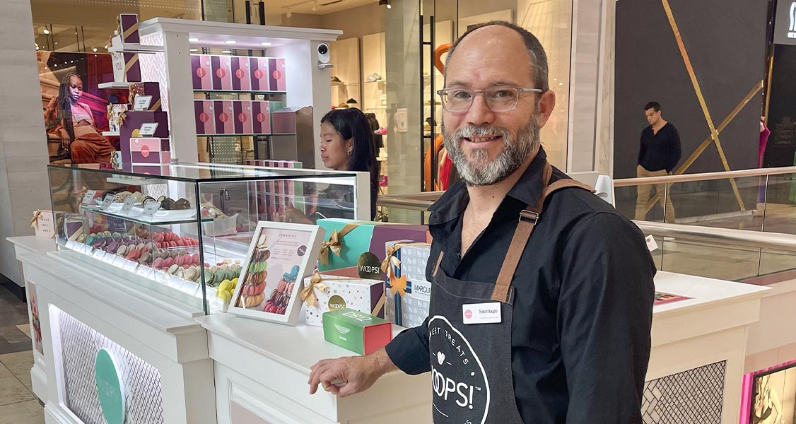 A smiling man with a Woops! apron is looking at the camera. His hand is resting on Woops! Macaron kiosk.