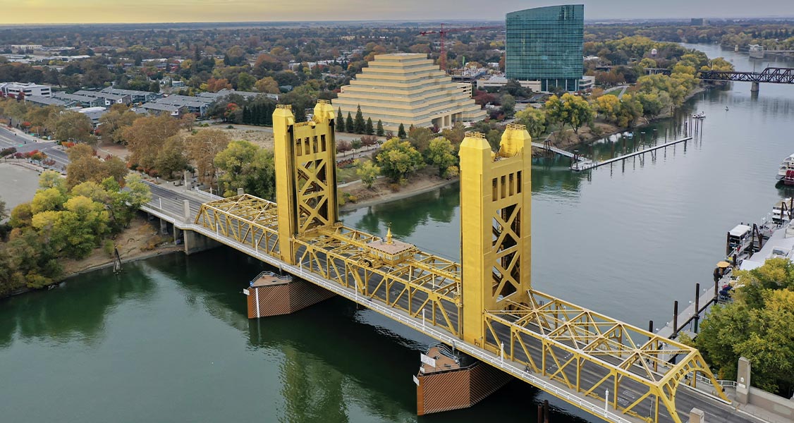 A yellow bridge over a wide river. Surrounding the bridge are houses and trees.
