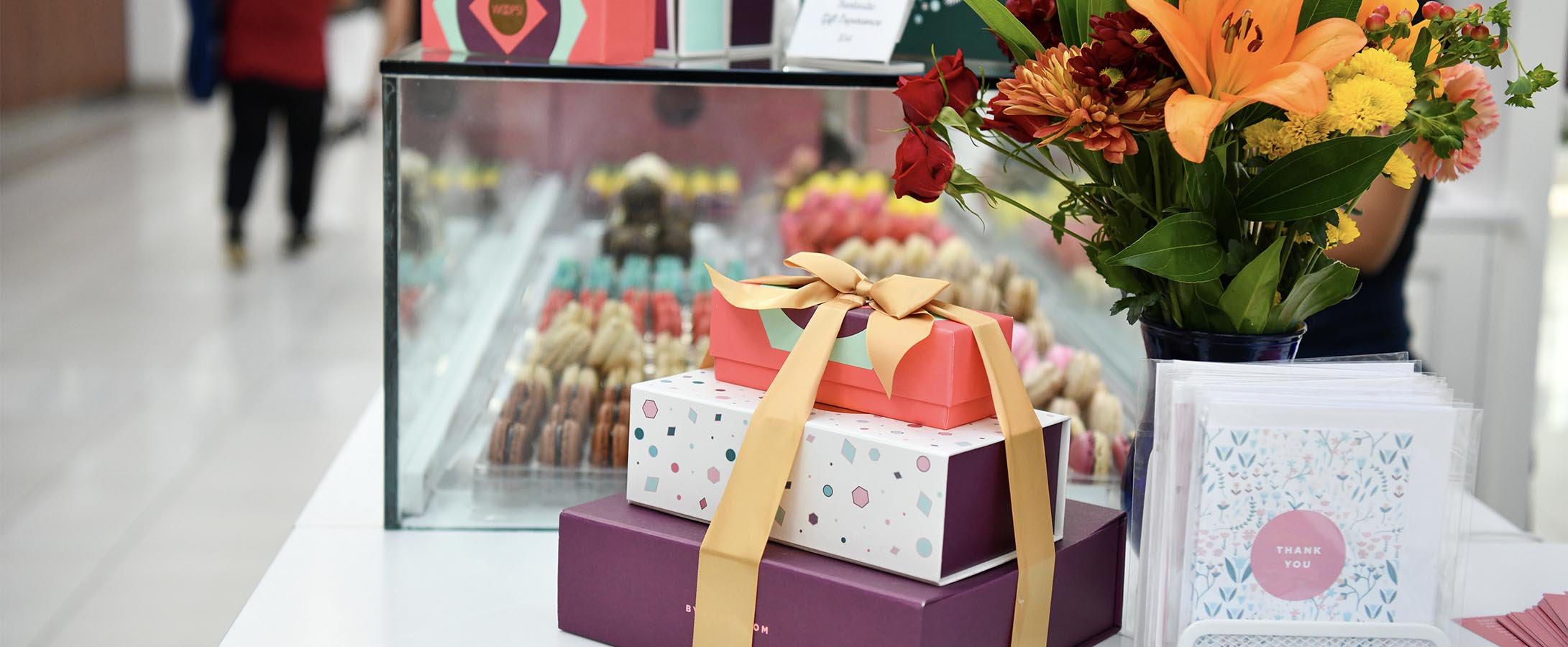A French macaron stack with a golden ribbon is on top of a white counter. To the side are some greeting cards and flowers. Behind a counter are French macarons and macaron boxes.