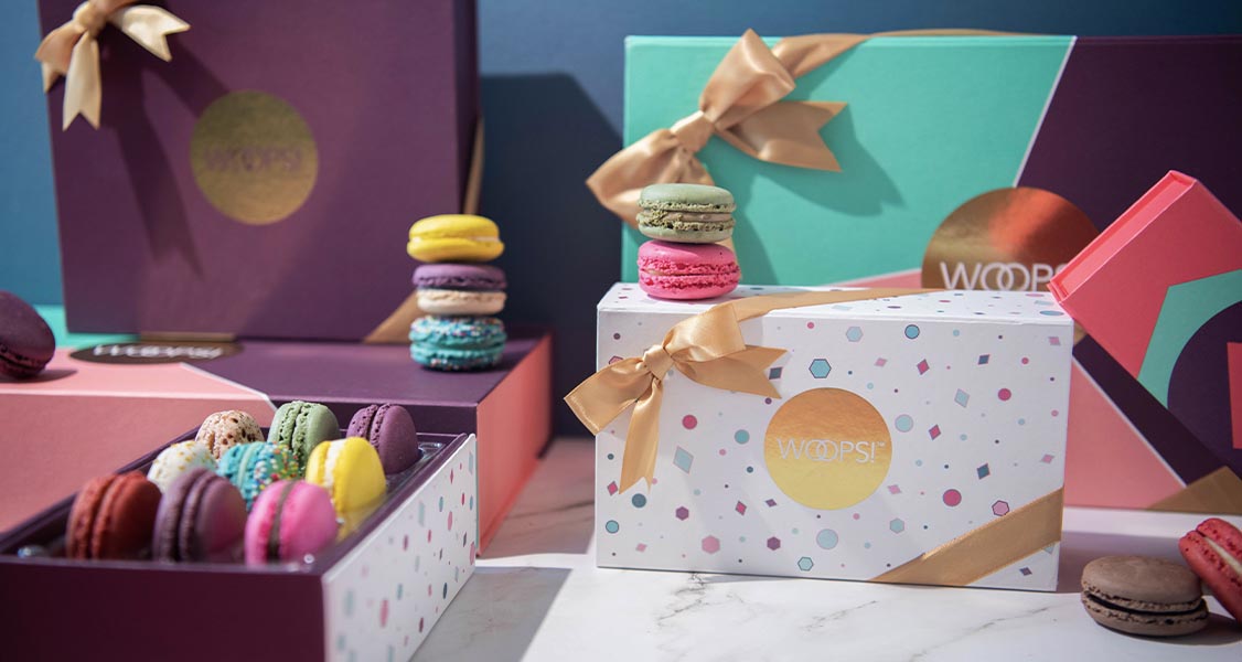 Numerous Woops! French macaron boxes with different custom sleeves and golden ribbons.