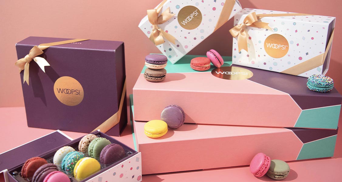 Several macaron boxes with golden ribbons are surrounded by assorted French macarons.
