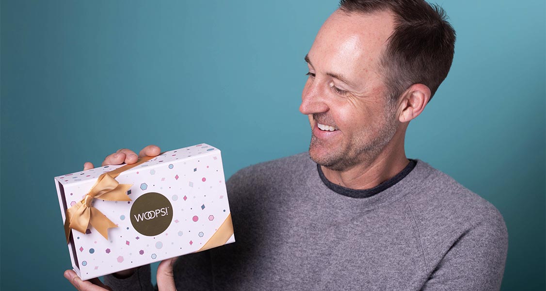 A smiling man with a grey t-shirt is holding a Woops! Macaron box with a golden ribbon.