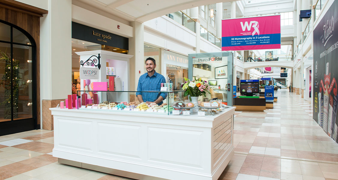 A smiling man is standing behind the counter of a Woops! Macarons & Gifts white kiosk. The kiosk is full of French macarons, macaron boxes, and flower vases.