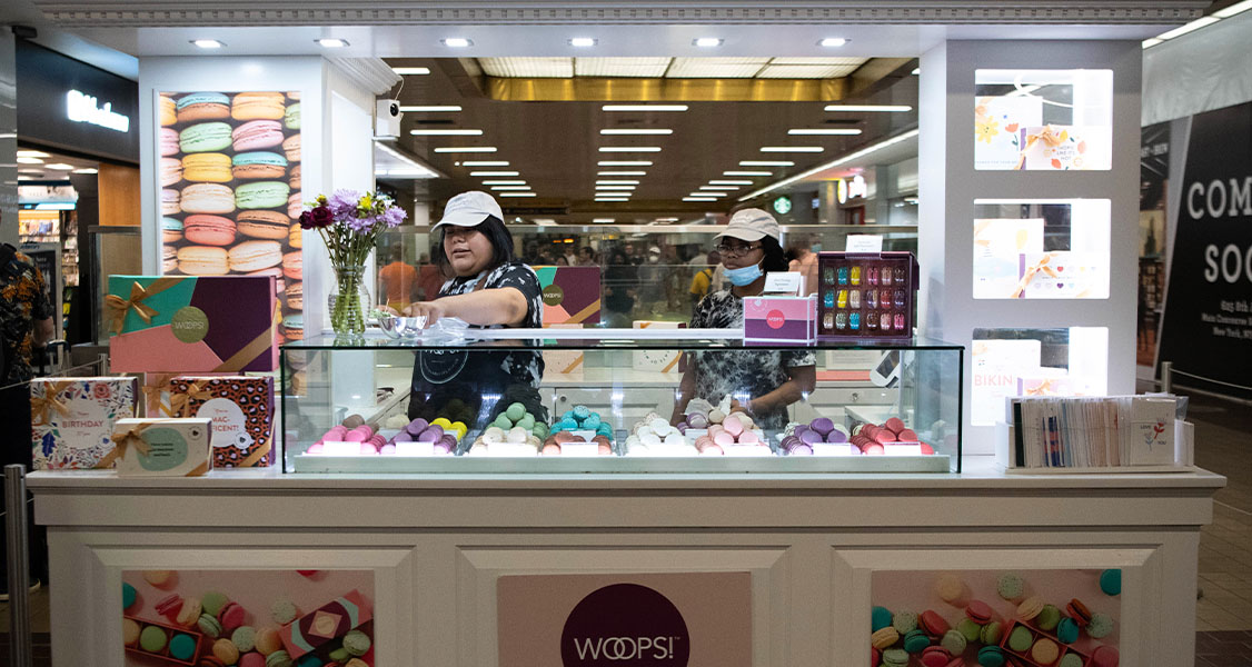 Two women are standing behind a Woops! Macarons & Gifts kiosk counter that’s full of assorted macarons and macaron boxes. To their sides are some marketing signs and people walking.