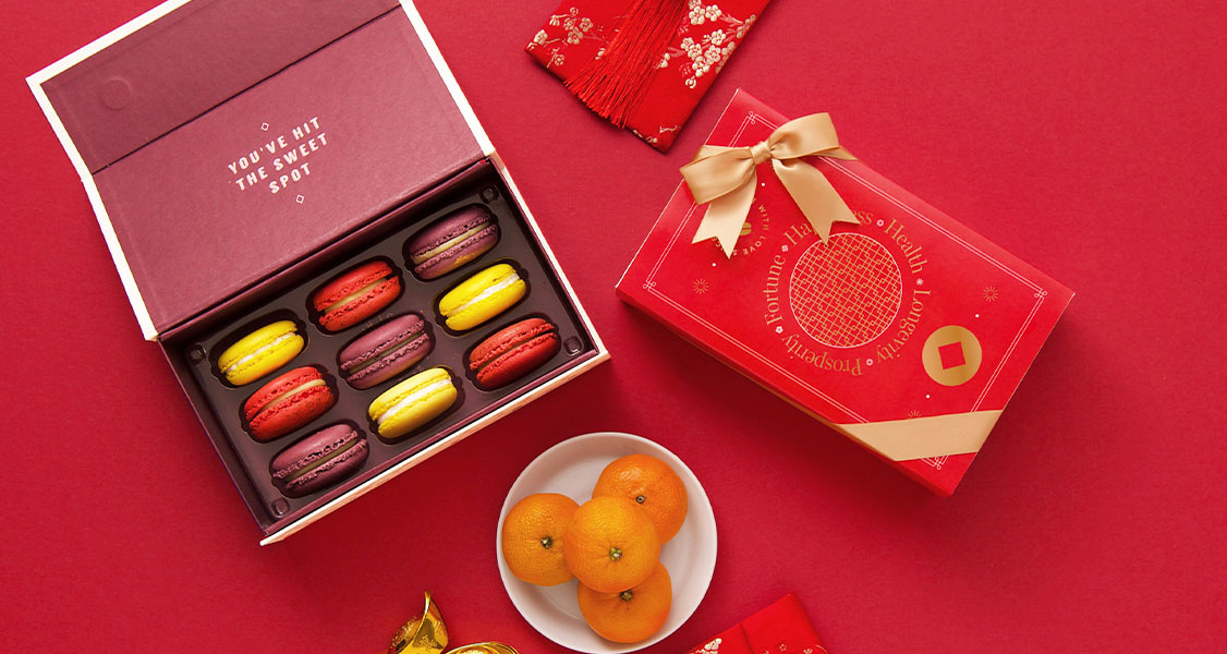 A box full of French macarons has a macaron box with a Lunar New Year sleeve to the left. Around them are some tangerines.