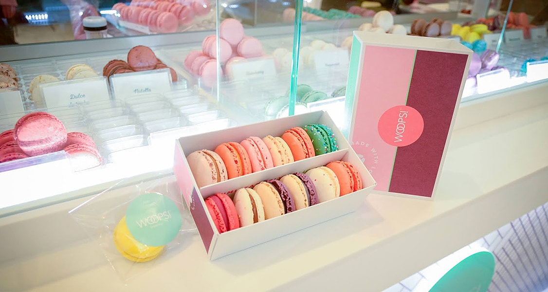 A box full of French macarons has a Woops! Pink macaron box to the right. Behind them is a counter full of assorted macarons.