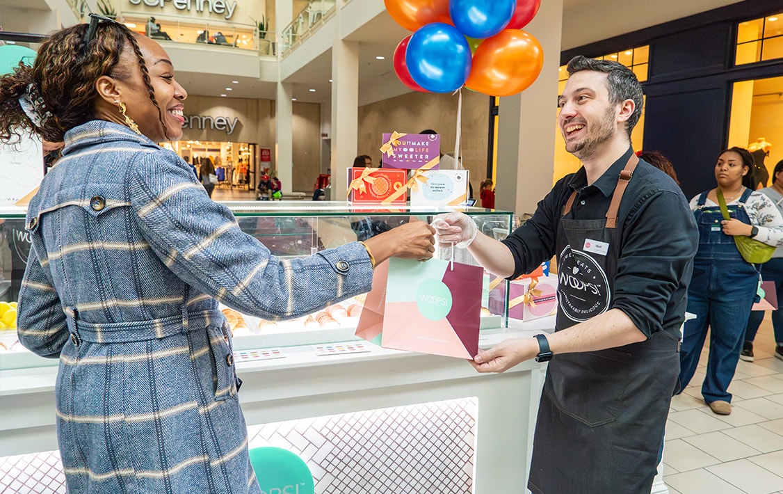 A smiling man with a Woops! Macarons & Gifts apron is giving a Woops! Gift bag to a smiling woman. Behind them is a Woops! Macaron kiosk.