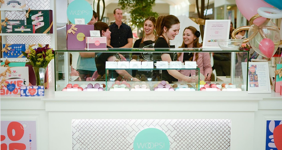 Several smiling women are behind a Woops! Macarons & Gifts counter full of macaron boxes and assorted macarons.