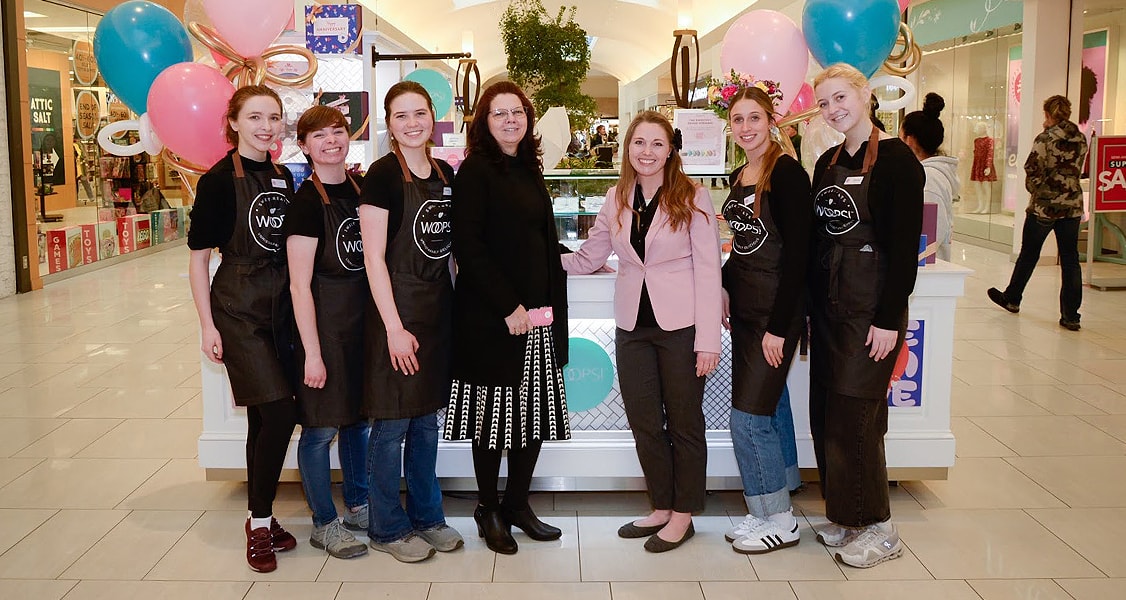 Seven smiling women are standing in front of a Woops! Macarons & Gifts kiosk decorated with pink and blue balloons.