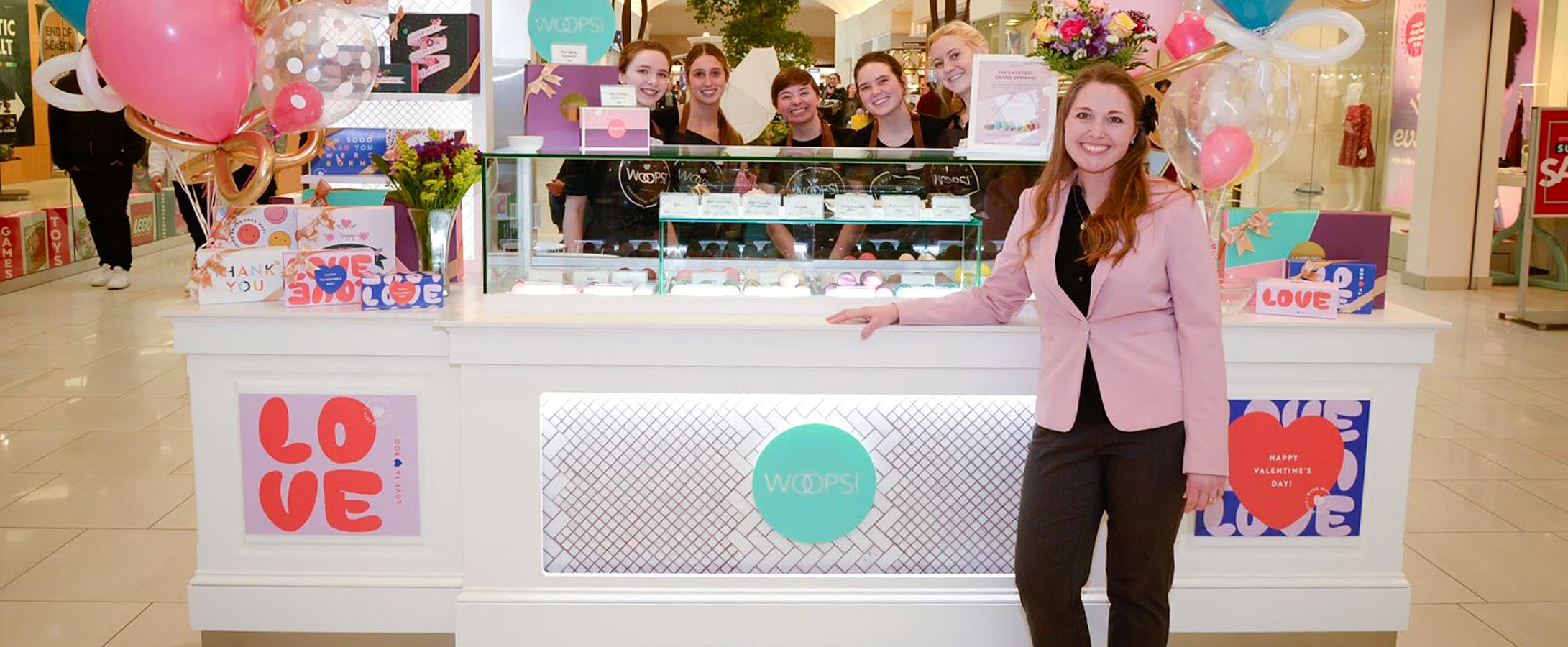 A woman standing in front of a Woops! Kiosk full of assorted macarons, balloons, and macaron boxes. Behind the counter are multiple smiling women.