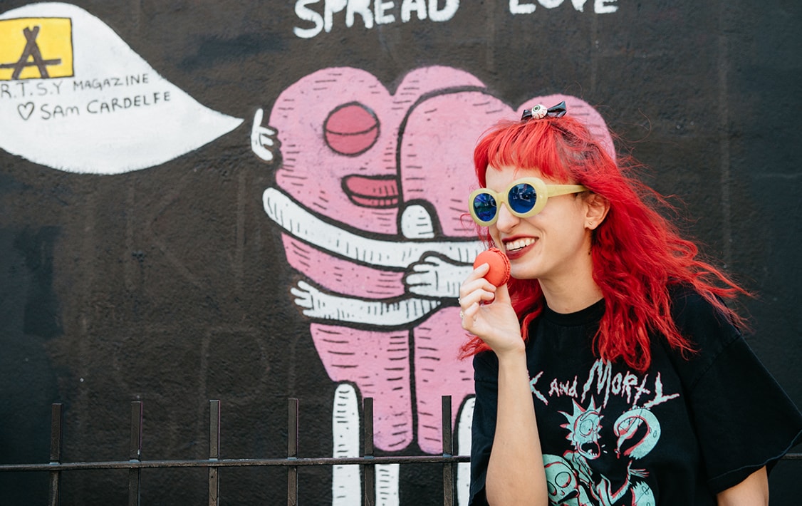: A woman with bright red hair and glasses is holding a Red Velvet macaron in her hand. Behind her is a hugging-hearts graffiti.