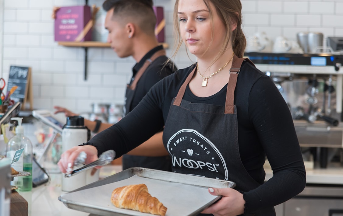 A woman with a Woops! Apron is carrying a baking tray with a croissant in her hands. Behind her is a man and surrounded by macaron boxes, coffee machines, and assorted sweets.