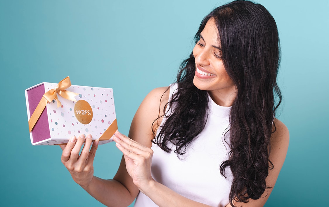 A smiling woman with dark hair is holding a Woops! Macaron box in her hands.