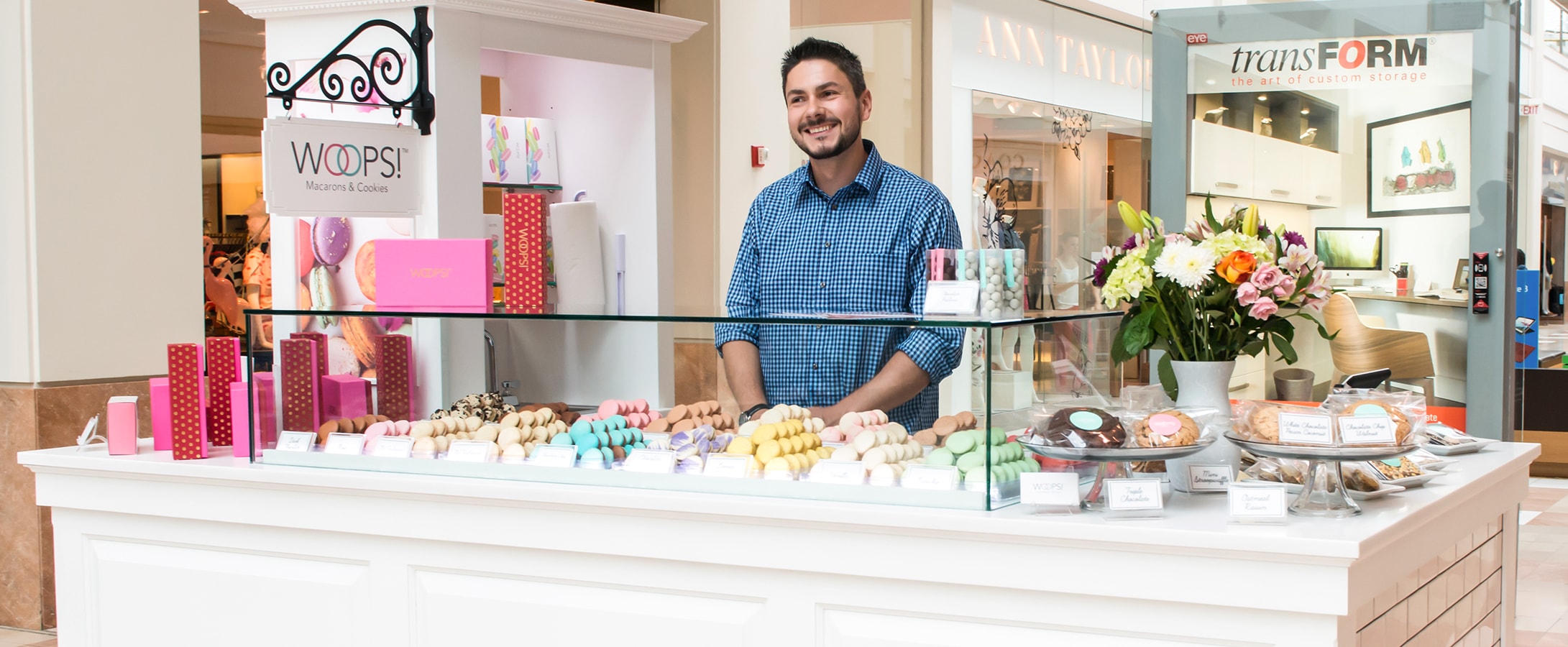 A smiling man is standing behind the counter of a Woops! Macarons & Gifts white kiosk. The kiosk is full of French macaron, macaron boxes, and flowers.