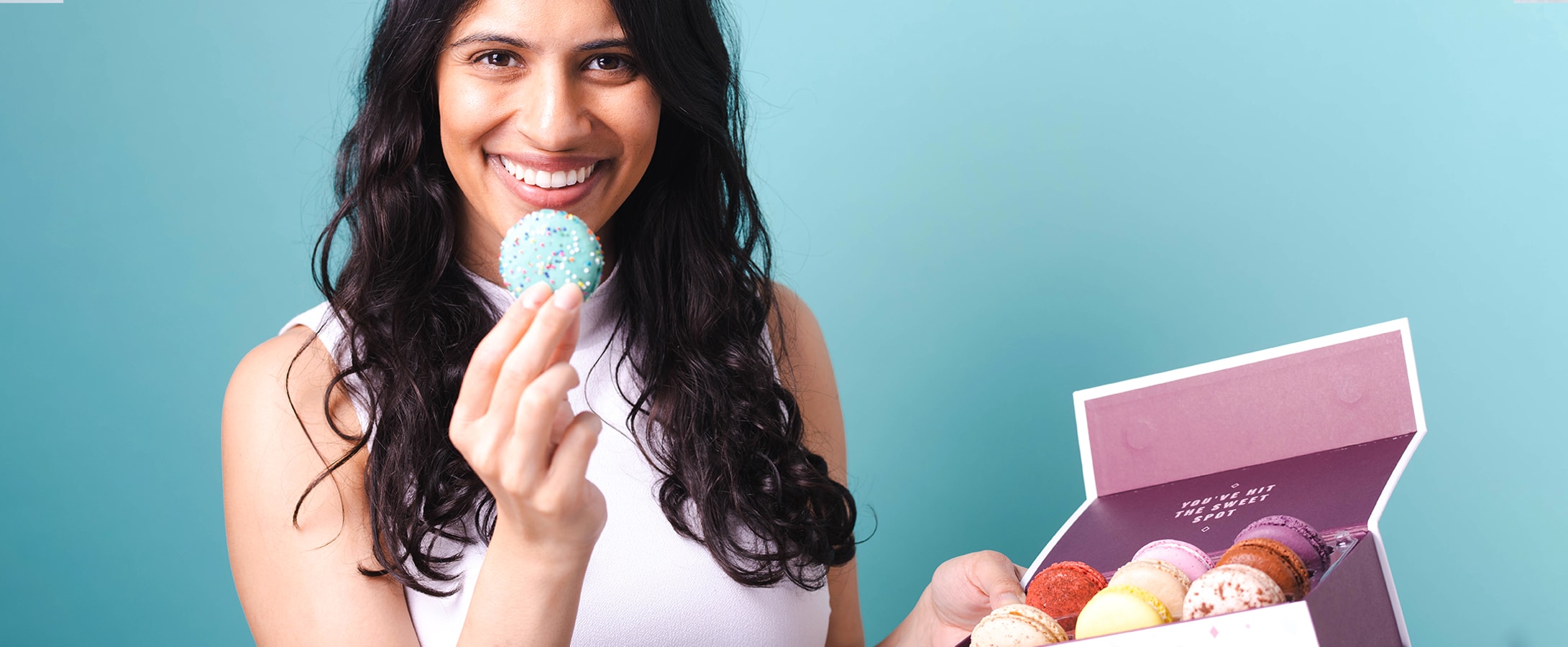 A smiling woman is holding a French macaron to her mouth and a box of macarons in the other.