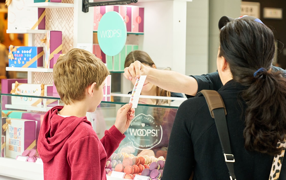 A kid holding a flavor guide in front of a Woops! Macarons & Gifts kiosk full of assorted macarons and macaron boxes.