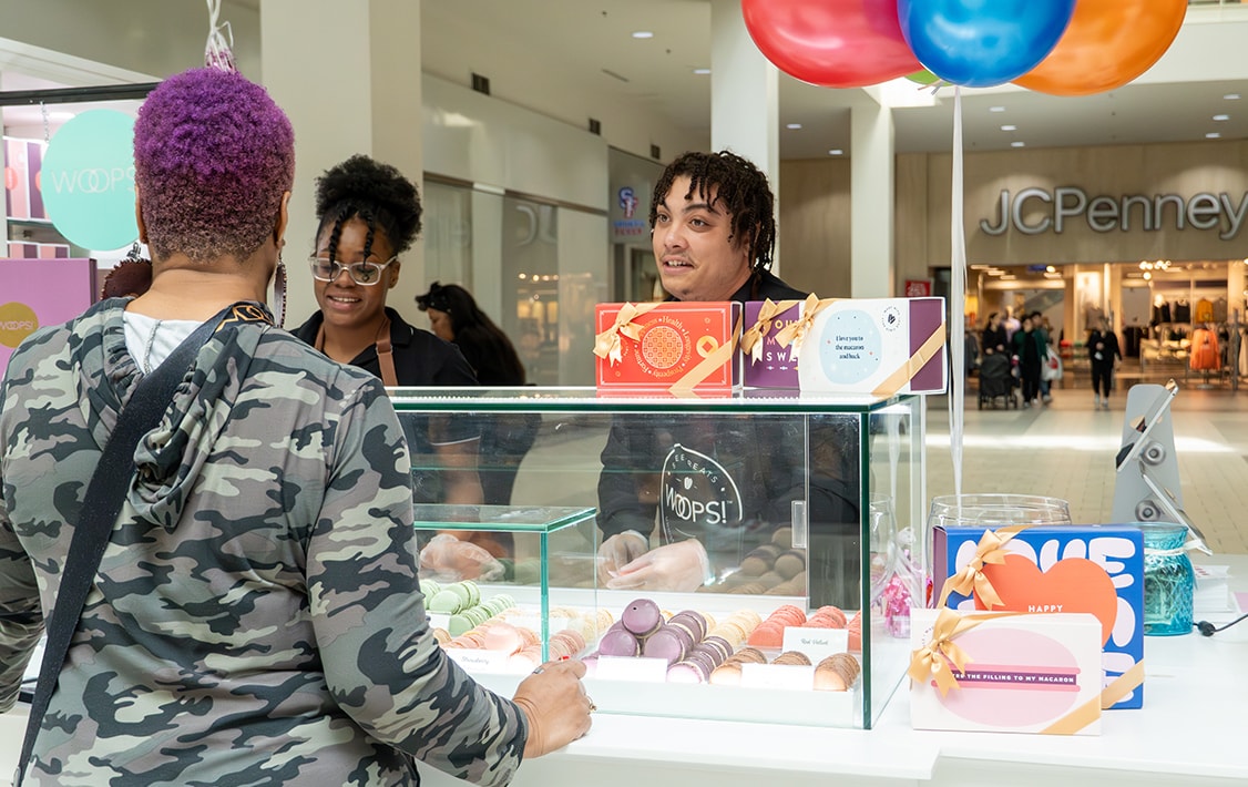 A man and woman behind a Woops! Macarons & Gifts kiosk counter is talking to a woman with purple hair.