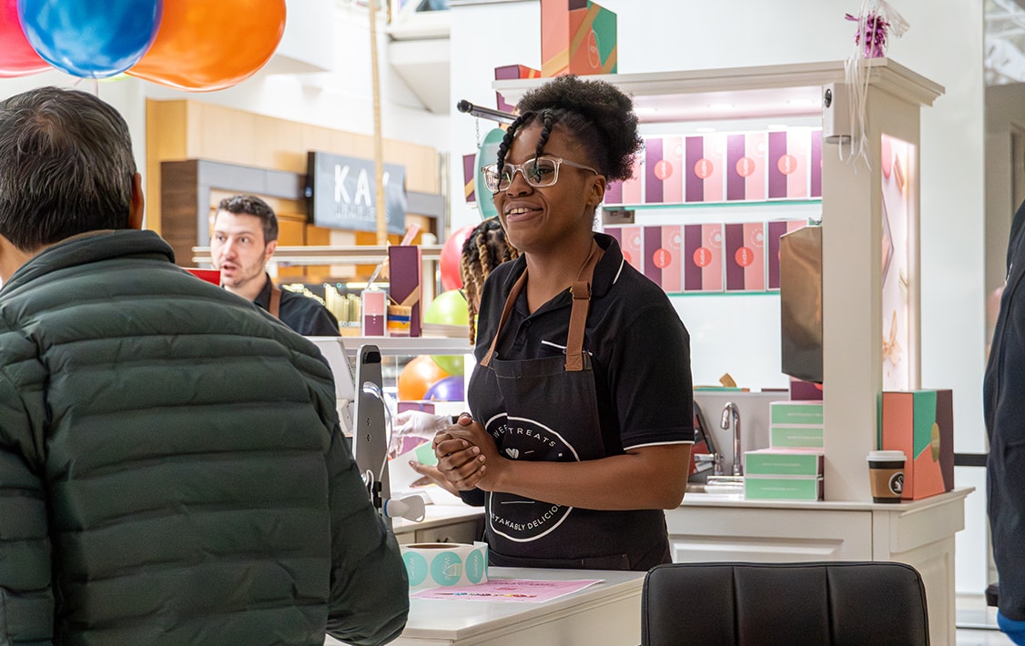 A smiling woman with glasses and a Woops! Apron is talking to a man. Behind her is a Woops! Macarons & Gifts kiosk counter full of macaron boxes.