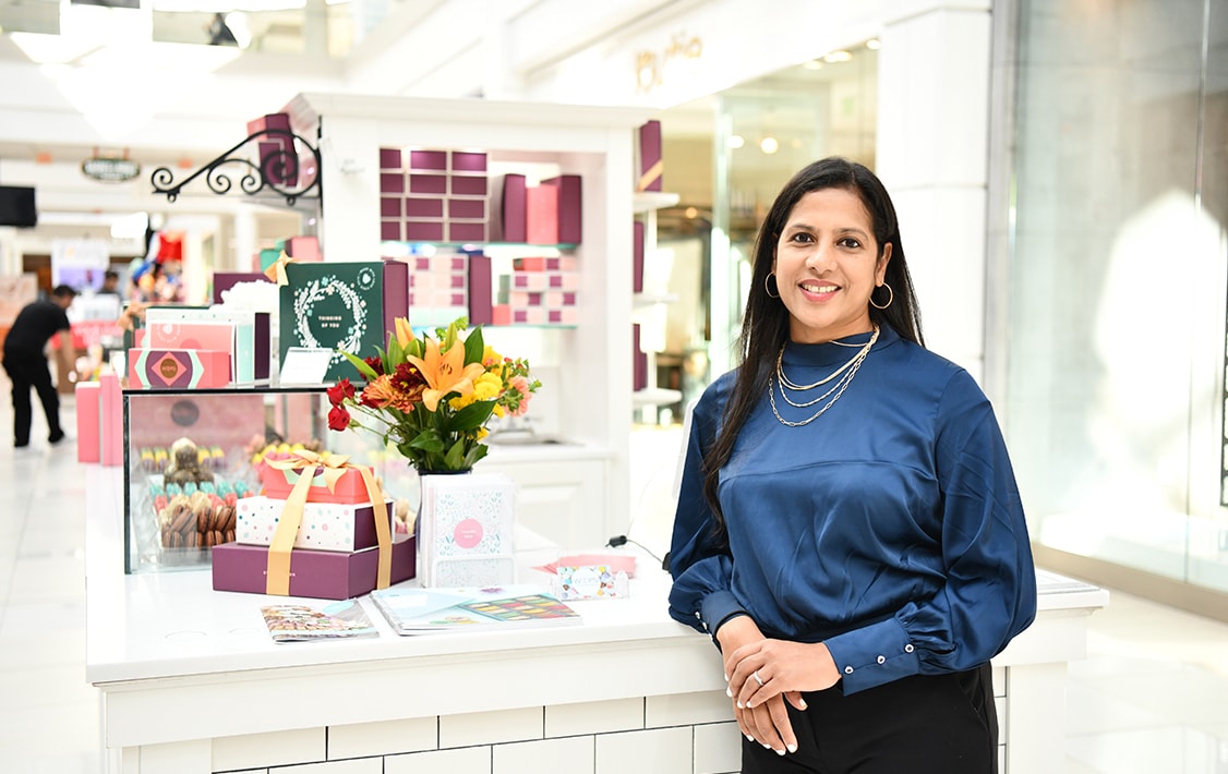 A smiling woman is leaning against the counter of a Woops! Macarons & Gifts kiosk full of assorted macarons and macaron boxes.