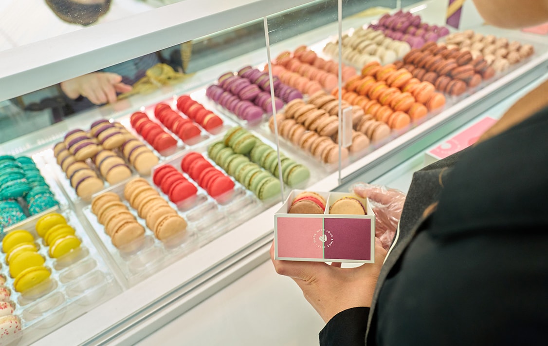 A woman is filling up a box with macarons. In front of her is a big Woops! macaron display in assorted flavors. 