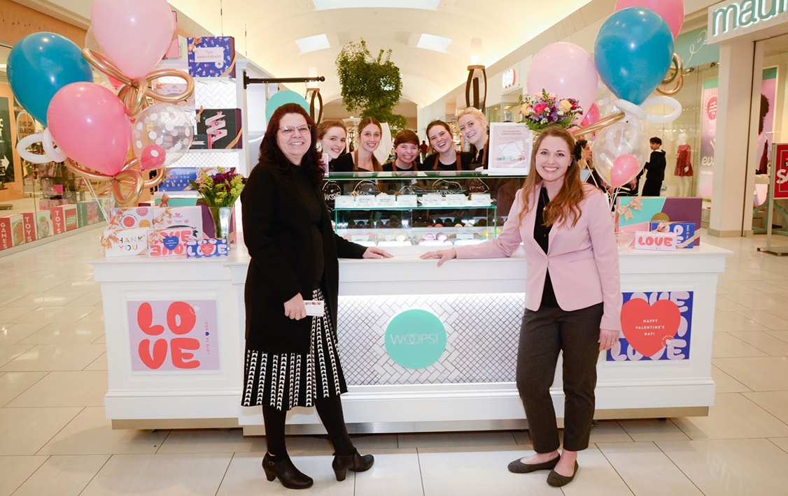 Two smiling women are standing in front of a Woops! Kiosk full of assorted macarons, macaron boxes, and balloons. Behind the counter are several smiling faces.