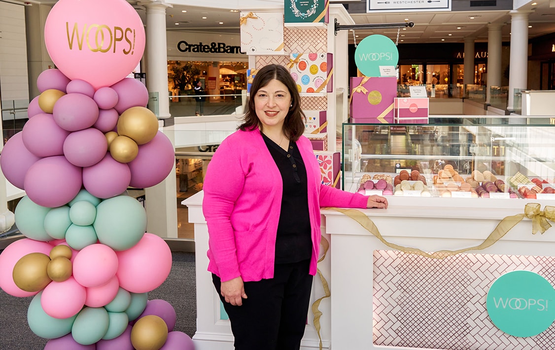  A smiling woman is standing in front of a Woops! Kiosk full of assorted macarons, macaron boxes, and colorful balloons.